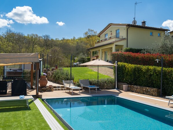 Une piscine dotée de chaises longues et d’un espace salon couvert, le tout entouré d’un jardin aménagé et d’une maison avec balcon.