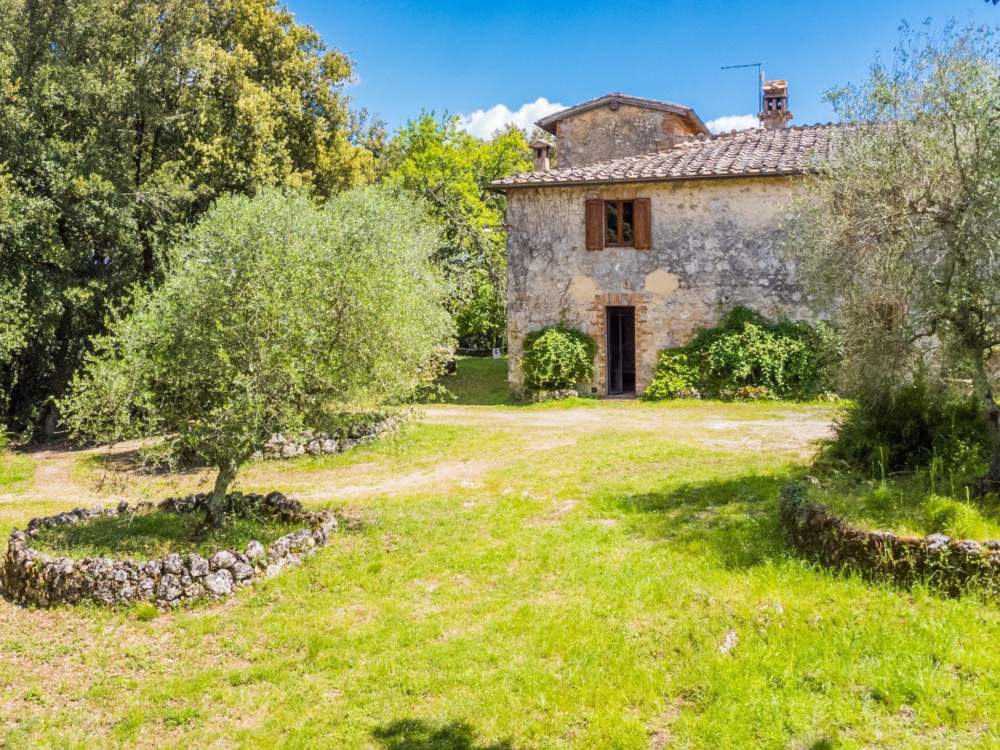 A rustic stone house with a tiled roof, surrounded by a well-maintained garden with trees and a stone wall.