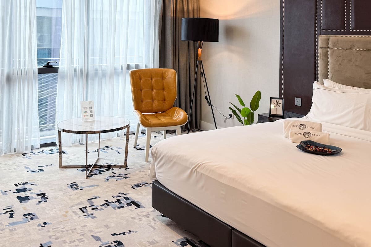 Ein Hotelzimmer mit einem großen Bett, einem kleinen runden Tisch, einem Ledersessel, einer Stehlampe und Blick auf die Stadt durch transparente Vorhänge.