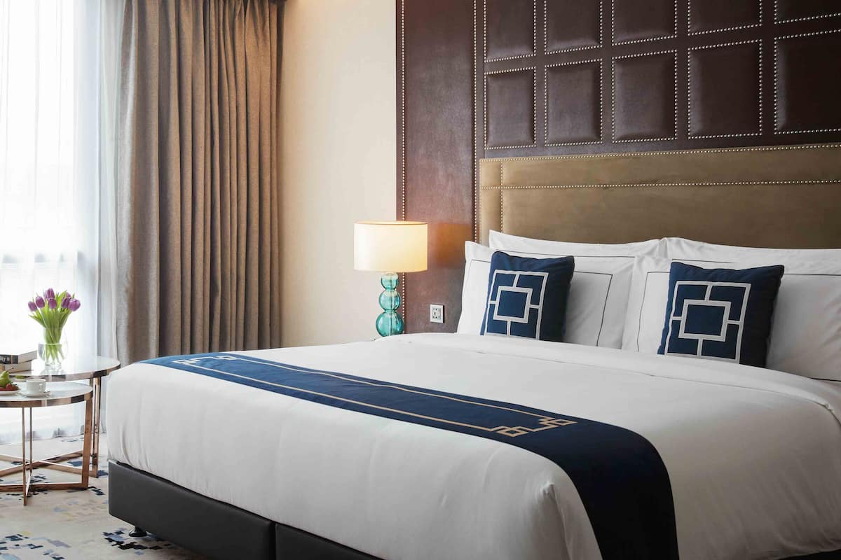 Ein Hotelzimmer mit einem großen Bett, einem Sessel, einem kleinen Tisch mit Lampe und Blick ins Freie durch transparente Vorhänge.