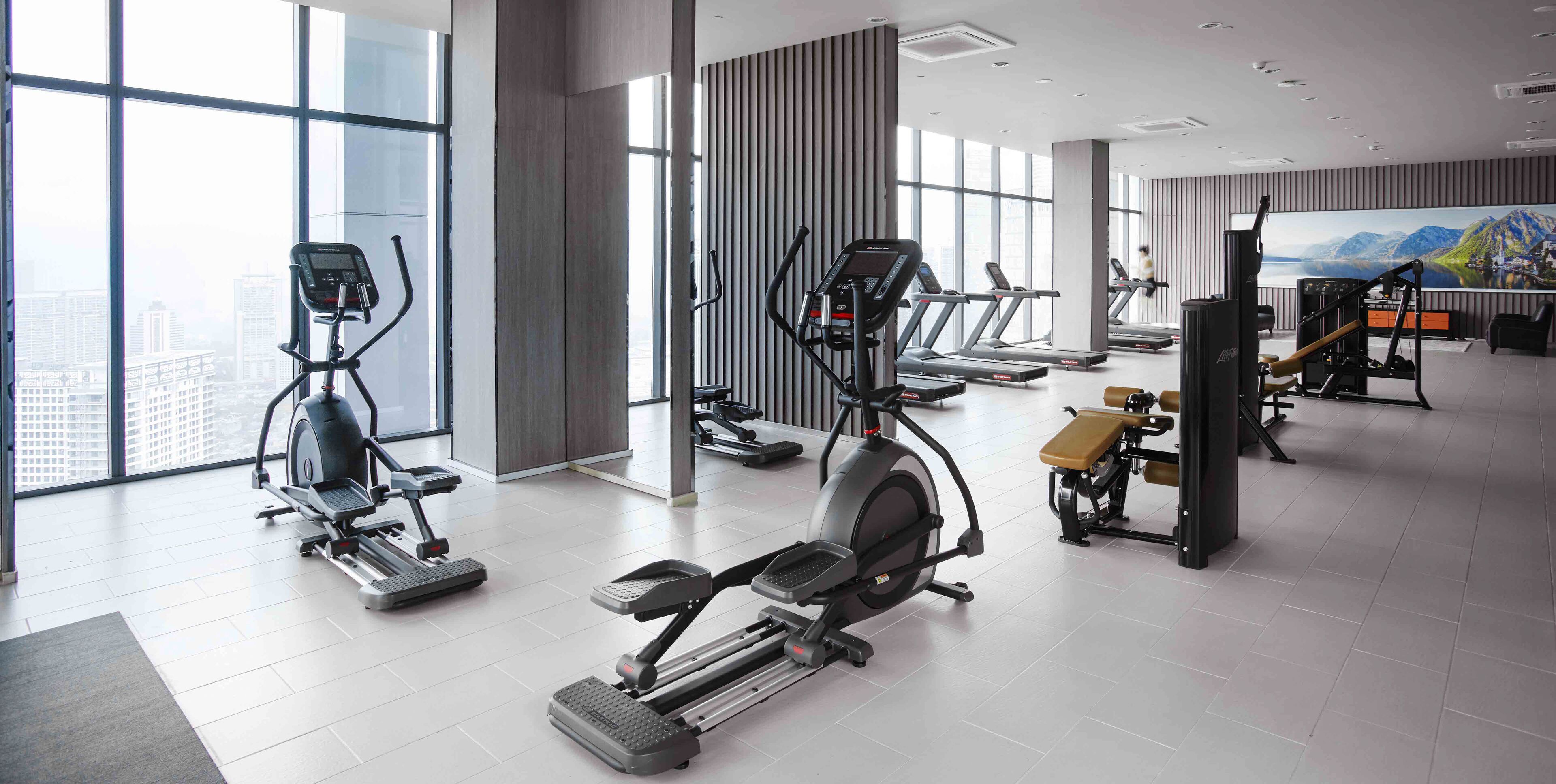 A modern gym with various cardio machines, large windows, and a cityscape view.