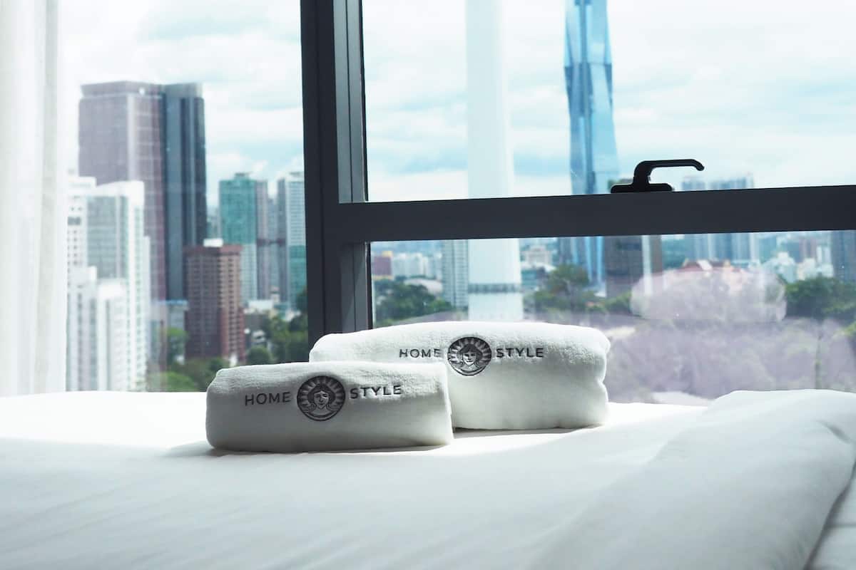 Ein Hotelzimmer mit einem großen Fenster, zwei Handtücher mit dem Schriftzug "HOME STYLE" und Blick auf eine Stadtlandschaft mit einem Turm und Wolkenkratzern.