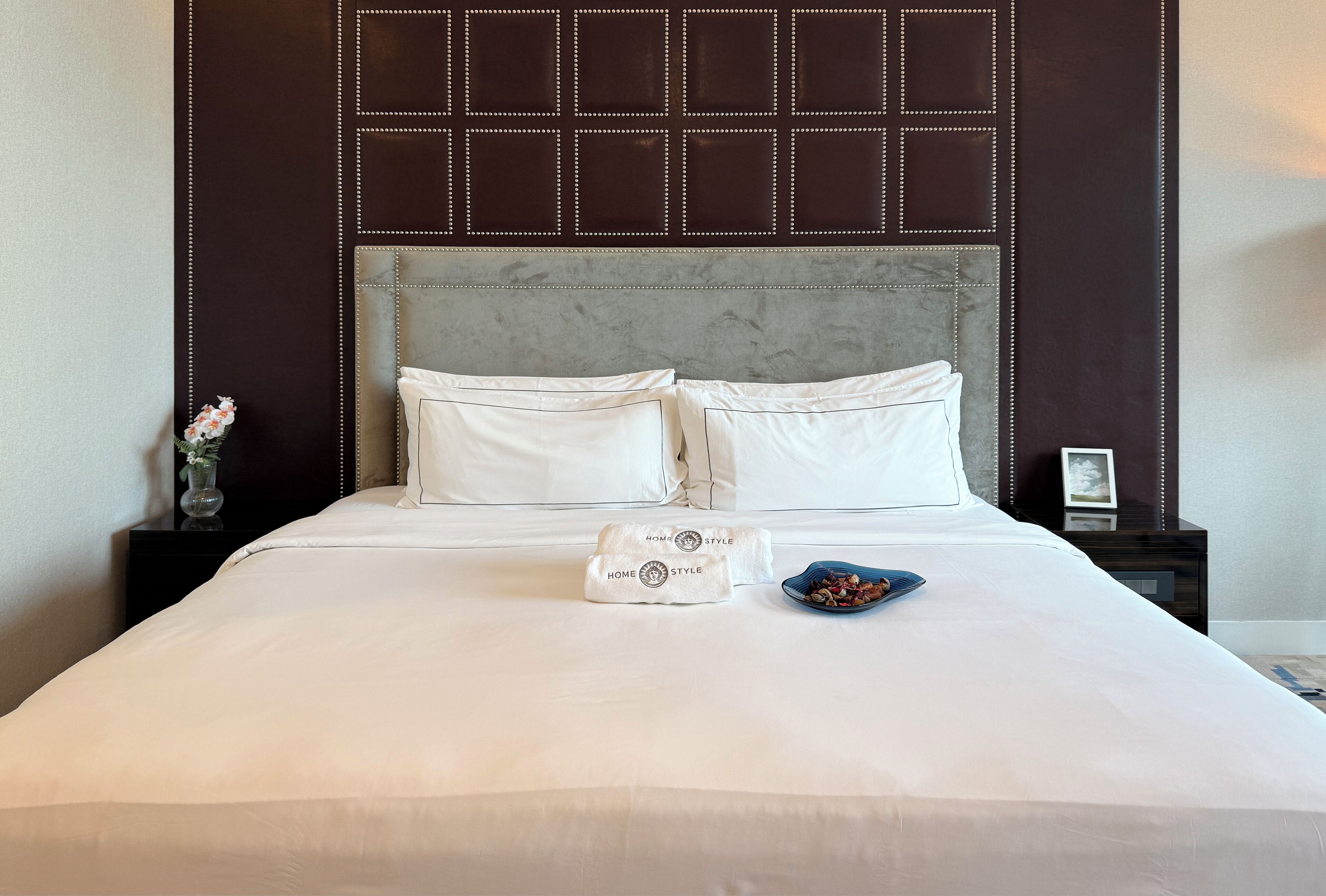 Una cama bien hecha con sábanas blancas, un cabecero de mármol y una mesita de noche con un florero y un marco de fotos.