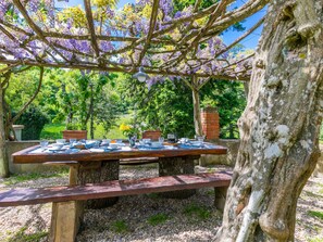 Uma área de refeições ao ar livre com uma mesa de madeira posta para quatro pessoas, cercada por árvores e um muro de tijolos.
