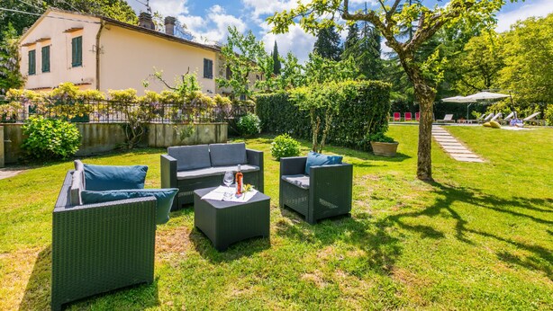 A patio with wicker furniture and a glass table, surrounded by a well-maintained lawn and a large tree.