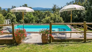 Une piscine rectangulaire avec une terrasse en bois et deux parasols blancs, entourée d’une végétation luxuriante et offrant une vue dégagée sur les collines lointaines.