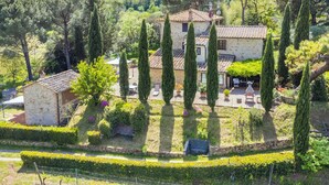 A stone villa with a tiled roof, surrounded by neatly trimmed hedges and tall cypress trees.