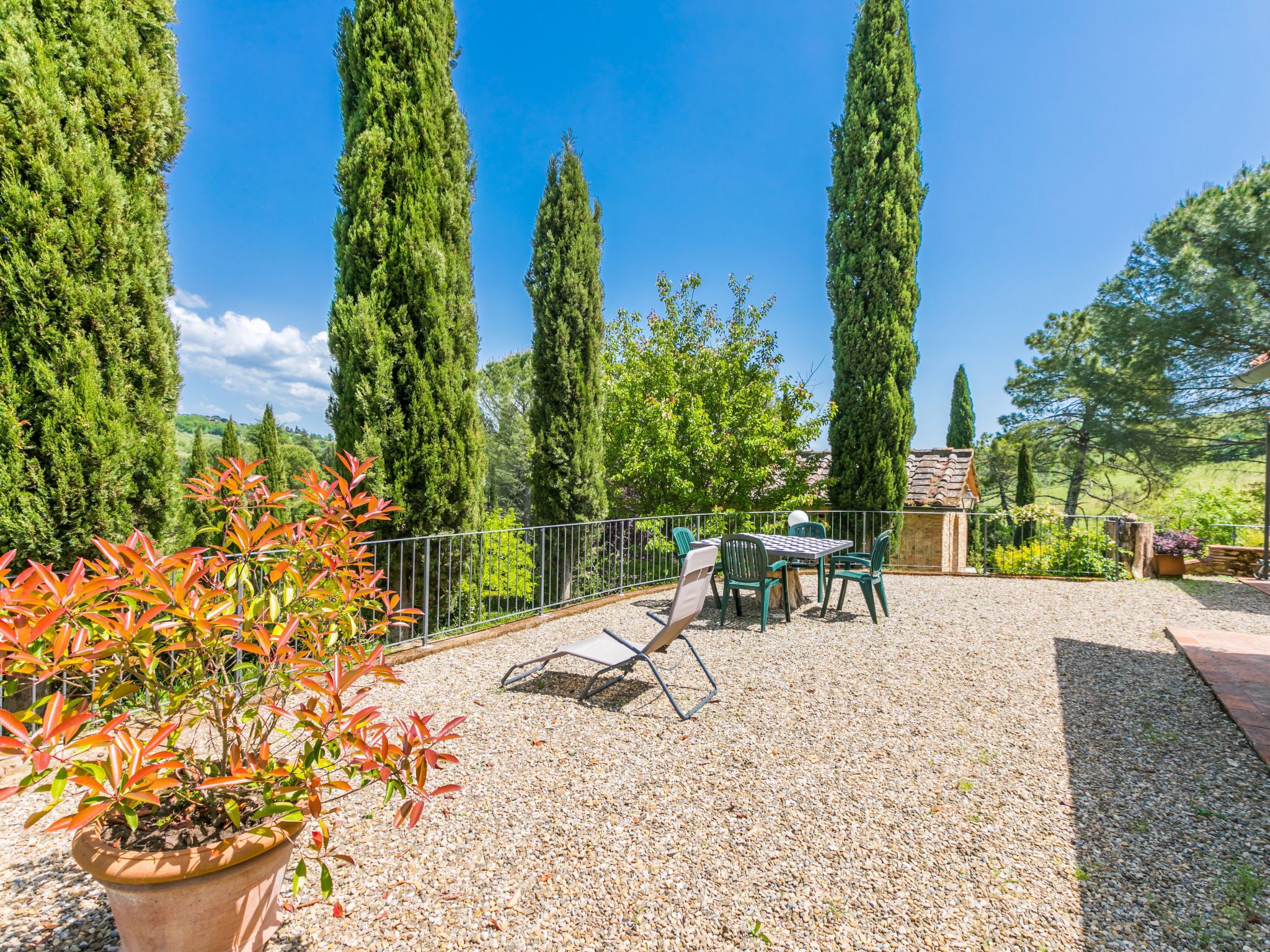A patio area with a table and chairs, surrounded by a gravel path and tall cypress trees.