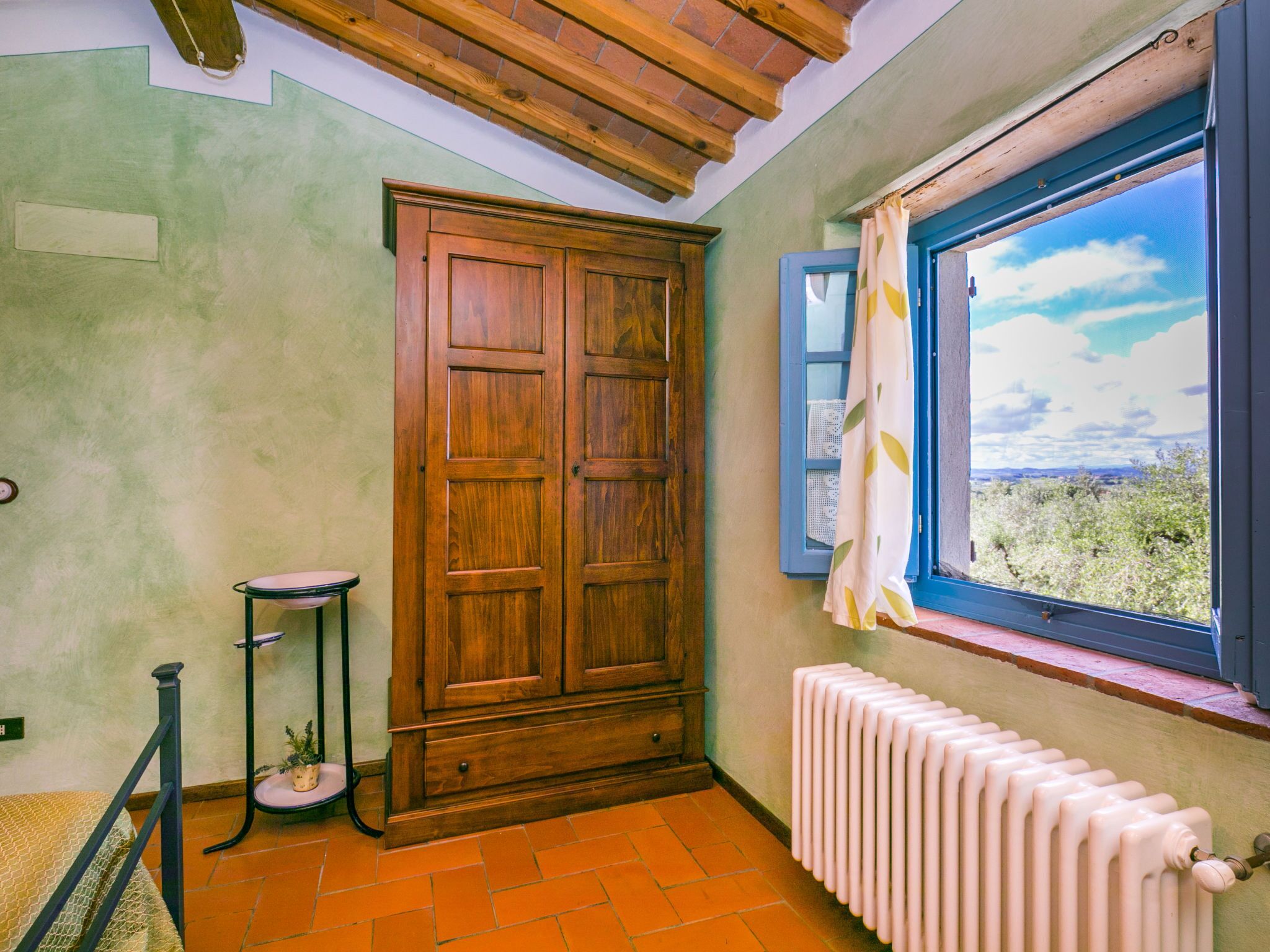 A room with a wooden wardrobe, a radiator, a window with a view, and a small table with a plant.