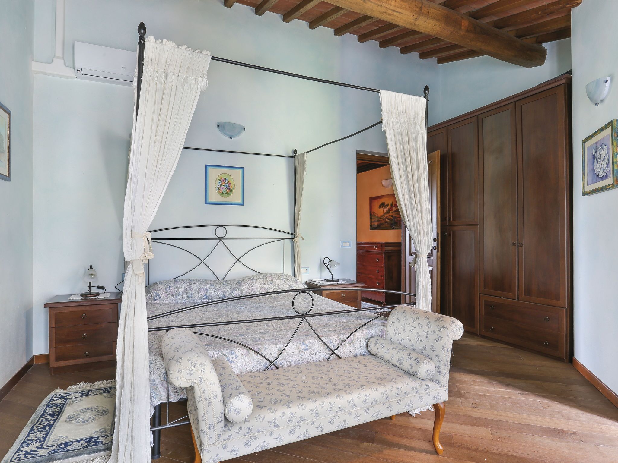 A four-poster canopy bed with a floral-patterned sofa, wooden flooring, and a wooden wardrobe.