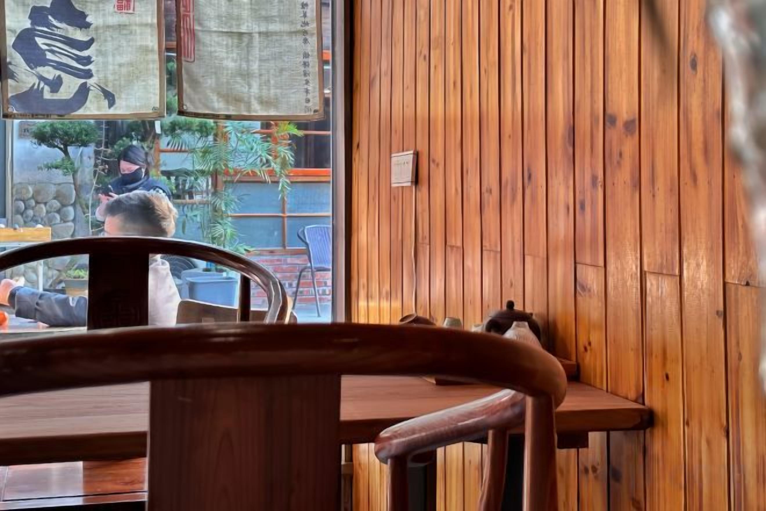 A wooden interior with a table and chairs, a window with a view of the outside, and a wall with a vertical wooden panel.