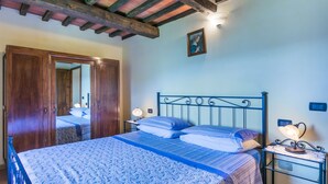 Un dormitorio con techo de madera, cama con colcha estampada azul, mesita de noche con lámpara y armario de madera.