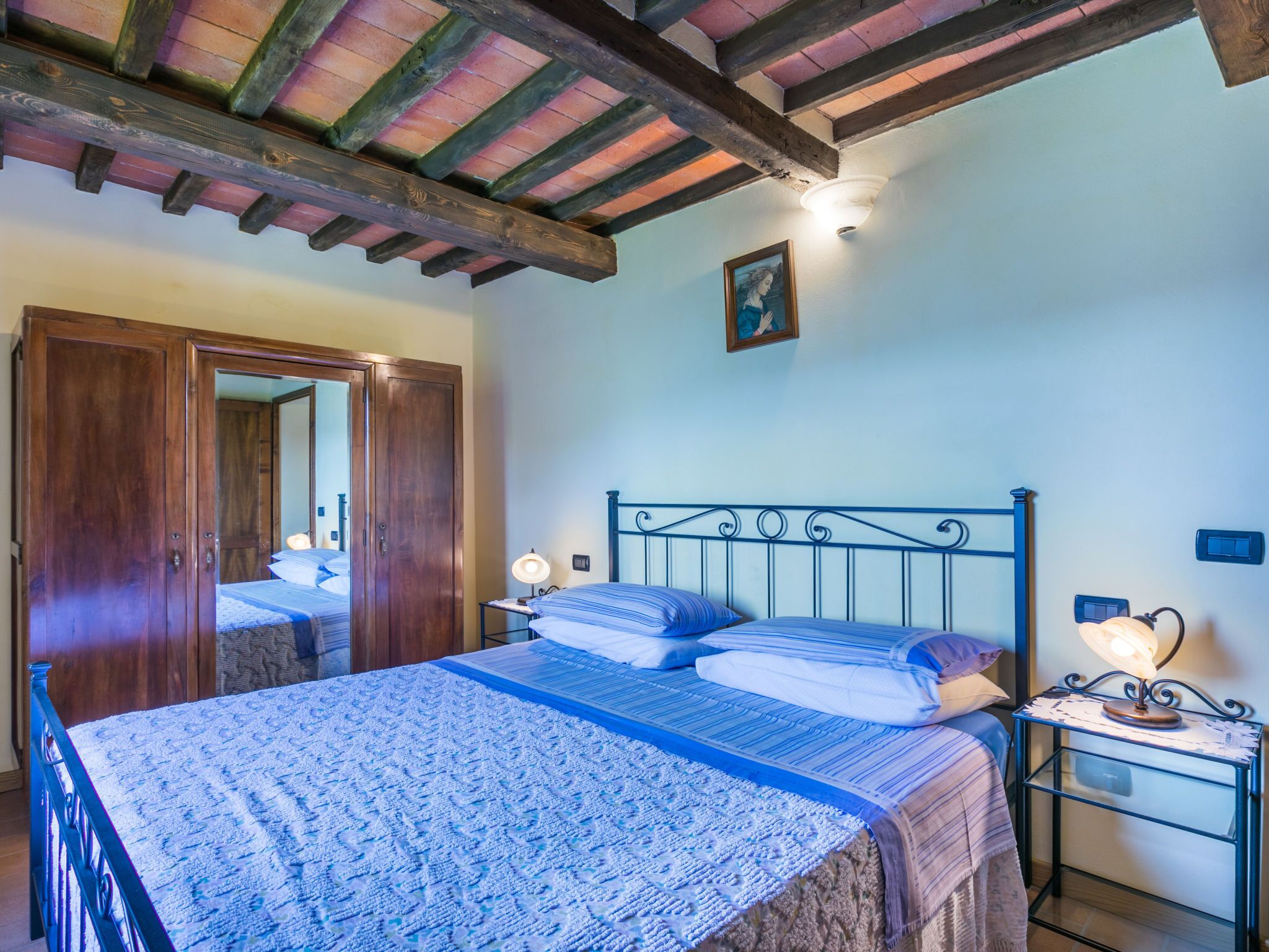Ein Schlafzimmer mit Holzbalkendecke, einem Bett mit blauen Bettbezügen mit Muster, einem Nachttisch mit Lampe und einem Holzschrank.