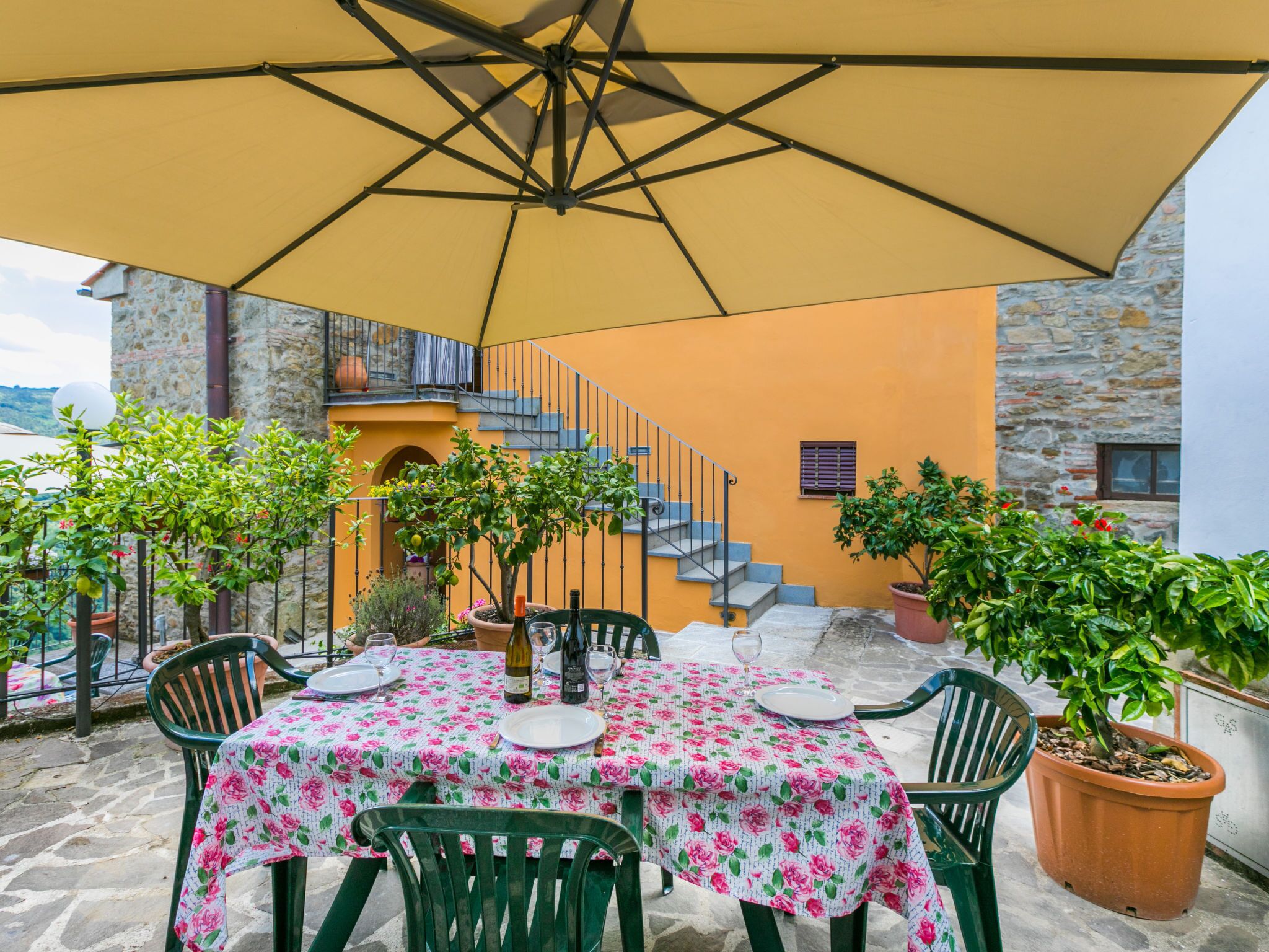 An outdoor patio with a table set for two, surrounded by potted plants and a yellow umbrella.