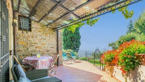 An outdoor patio with wicker furniture, a table with a patterned tablecloth, and a view of a garden and trees. - La Cupola: Geranio by Interhome (Pescia)