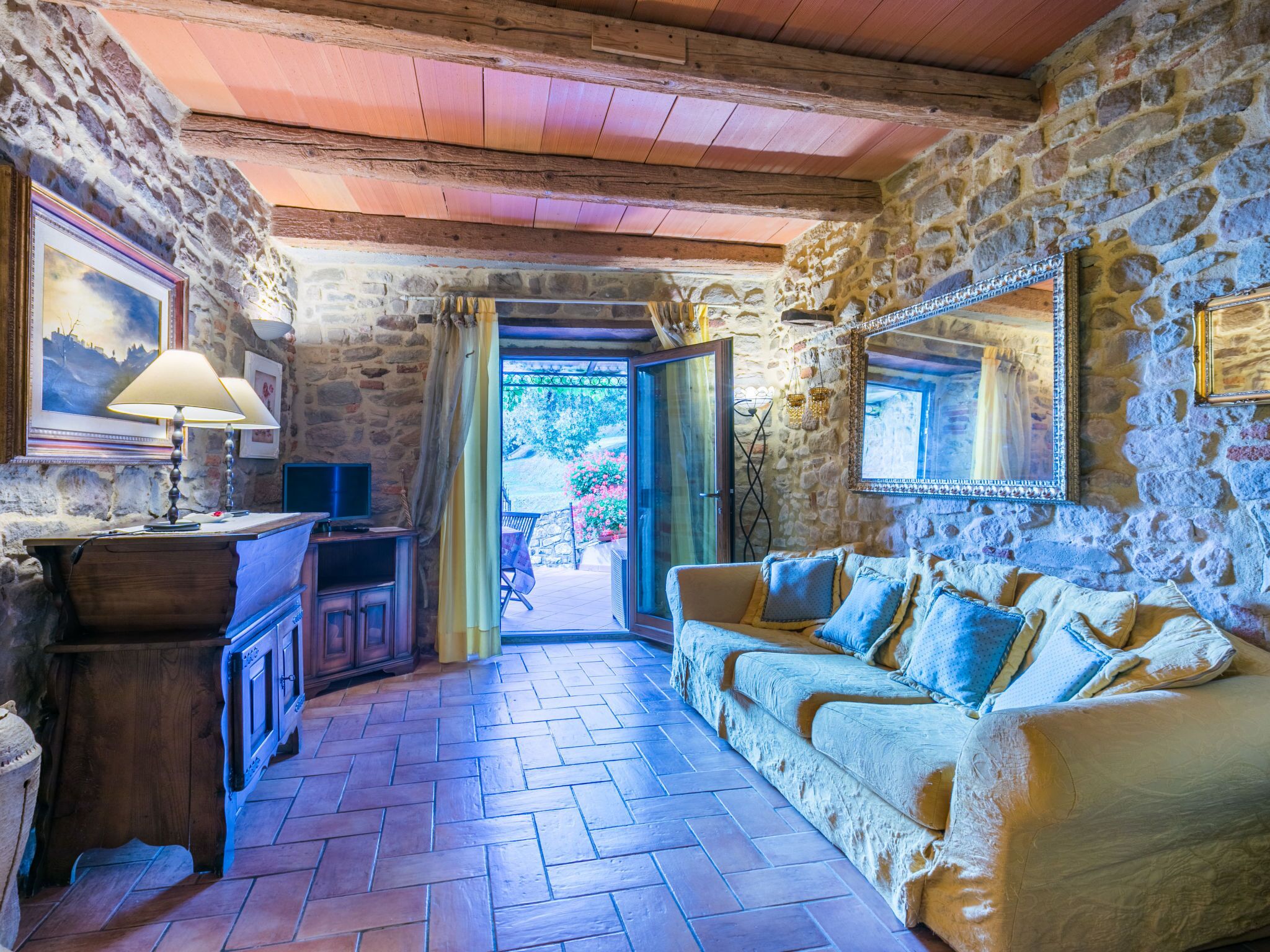 A rustic stone-walled room with a wooden ceiling, a sofa, a desk with a lamp, and a television.