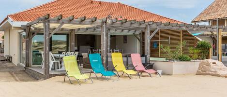 Un bâtiment situé en bord de mer, doté d’un patio couvert avec des fauteuils colorés, une pergola en bois et une plage de sable en premier plan.
