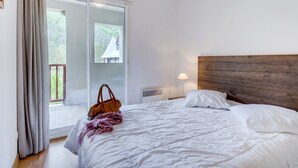 A bedroom with a large bed, a wooden headboard, a nightstand with a lamp, a window with a view of a house, and a sliding glass door leading to an outdoor area.