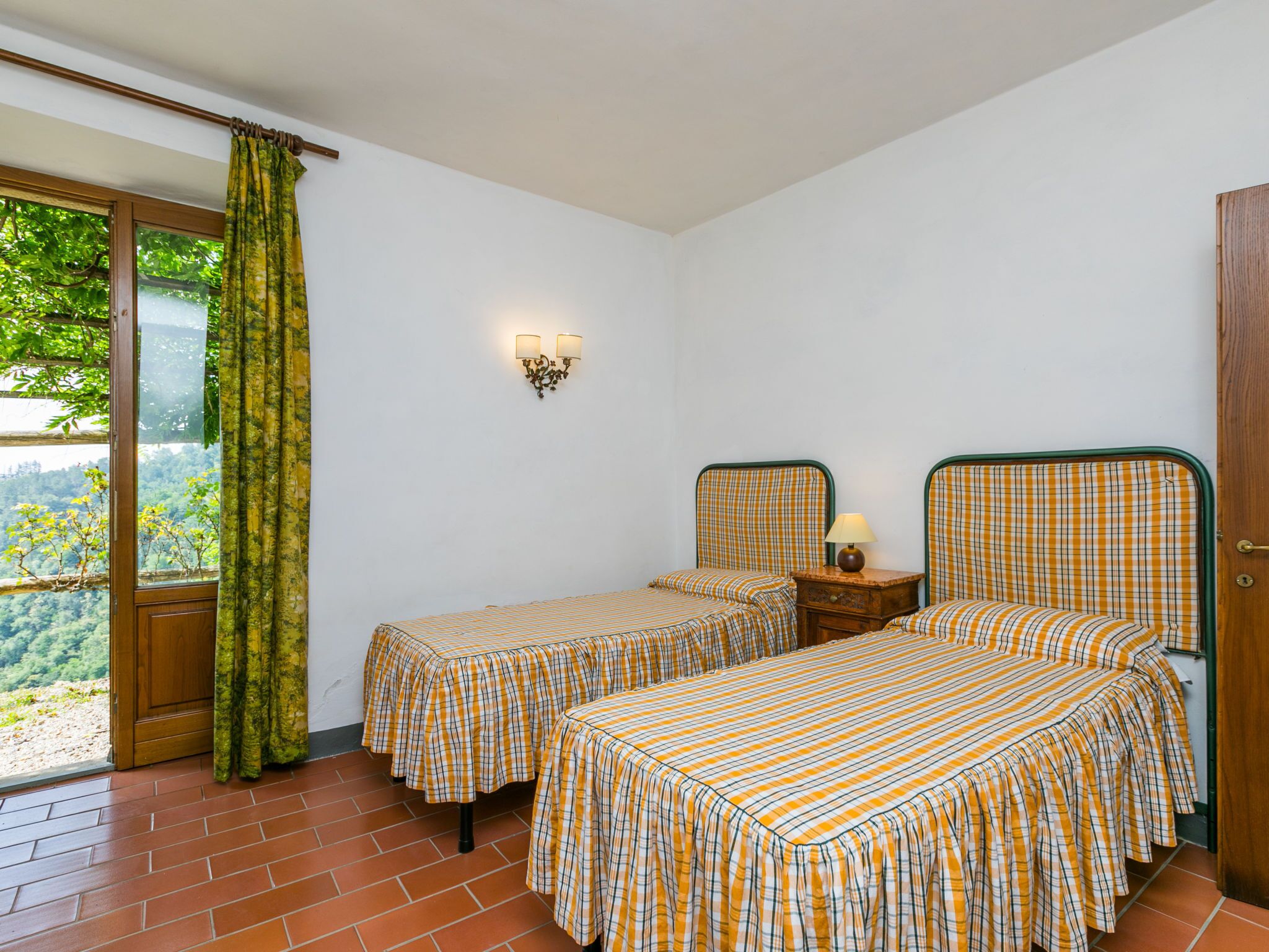 A room with two beds, a window with a view of greenery, a wall-mounted light fixture, and a door.