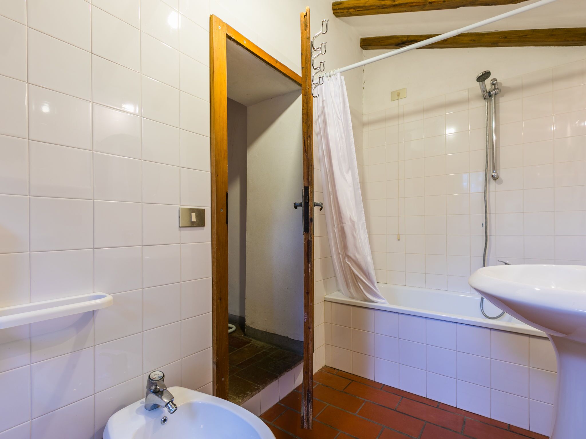 A bathroom with white tiles, a white sink, a shower with a curtain, and a wooden door frame.