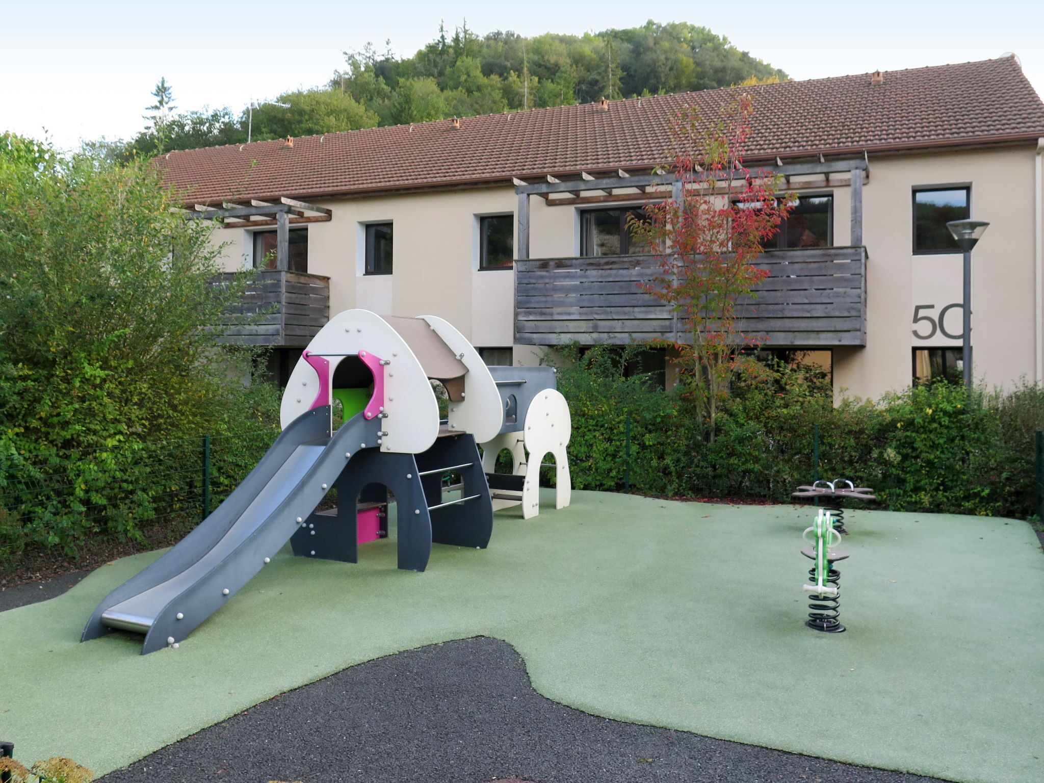 Un terrain de jeux avec un toboggan et des balançoires, entouré de verdure et bordé d’un bâtiment doté de balcons.