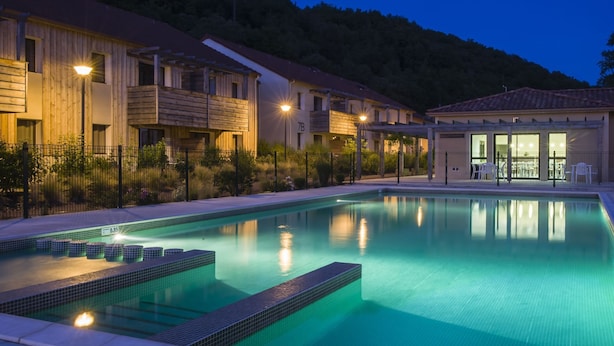 Una piscina con bordo piastrellato, circondata da edifici con facciata in legno e un'area coperta con tavoli e sedie.