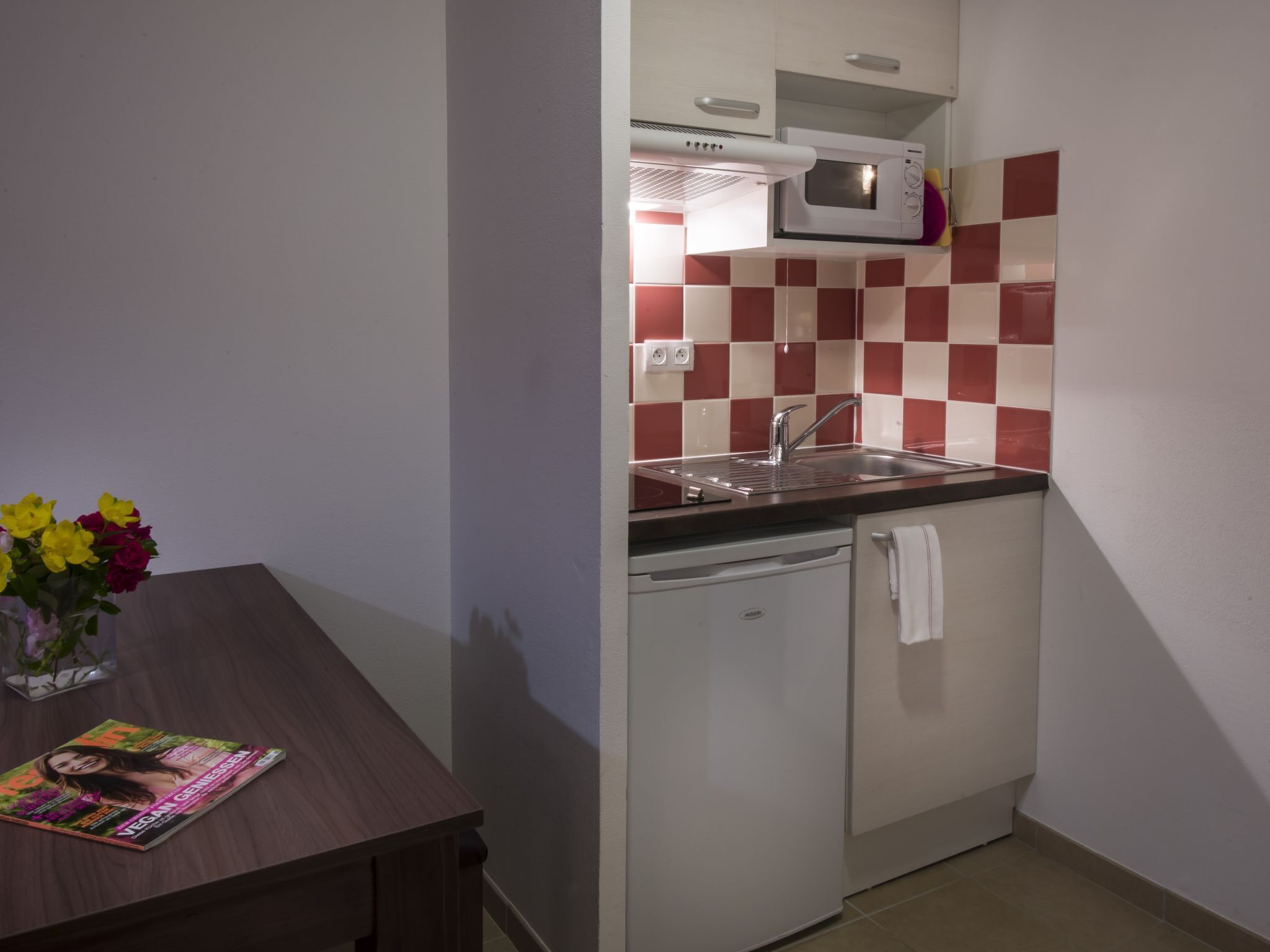 A compact kitchen with a microwave, sink, and dishwasher. A table with a magazine and flowers is visible.