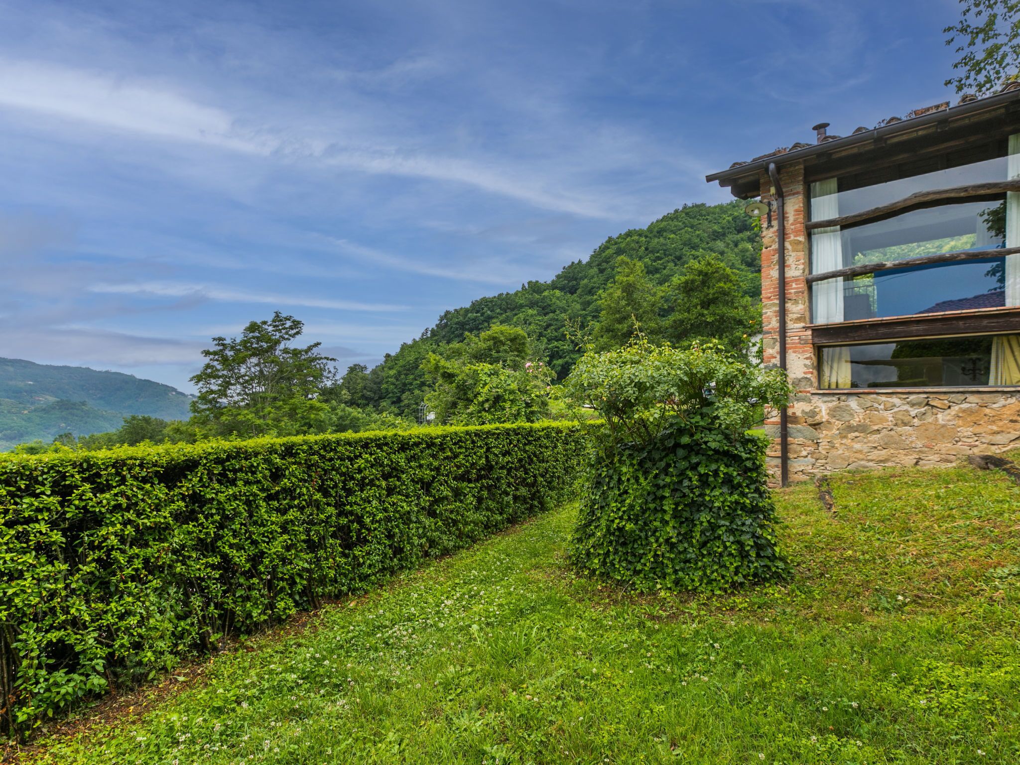 A stone building with a glass window, surrounded by lush greenery and hills in the background.