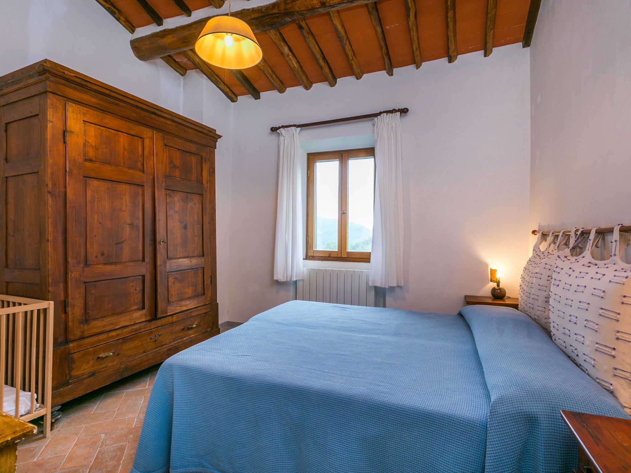 Un dormitorio con un armario grande de madera, una cama con cubrecama azul, una ventana con cortinas blancas y una mesita de noche con lámpara.