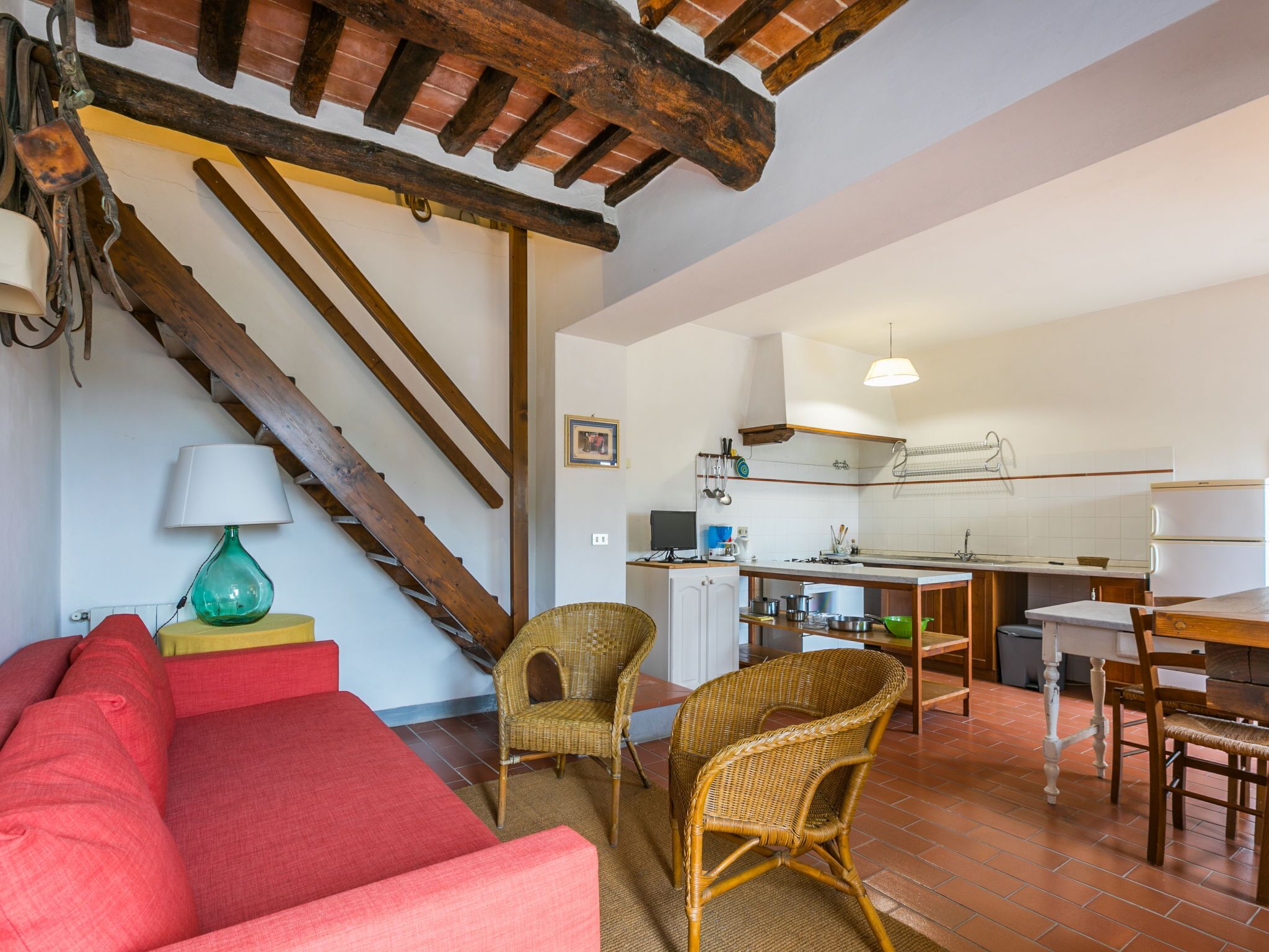 A spacious living area with a red sofa, wicker chairs, a wooden staircase, and a kitchen with white cabinets and a hanging pot rack.