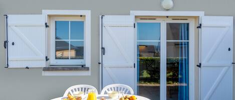 An outdoor dining area with a table set for breakfast, two windows with shutters, and a door with glass panels.