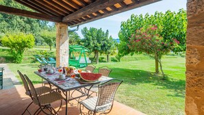 Une terrasse couverte avec une table dressée pour le repas, entourée de chaises, et une vue sur un jardin luxuriant, avec des arbres et un ciel dégagé.