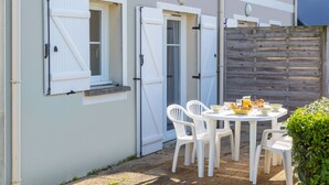Un patio con una mesa redonda y sillas de plástico blanco, cerca de una valla de madera, y un edificio con contraventanas blancas y un muro gris.