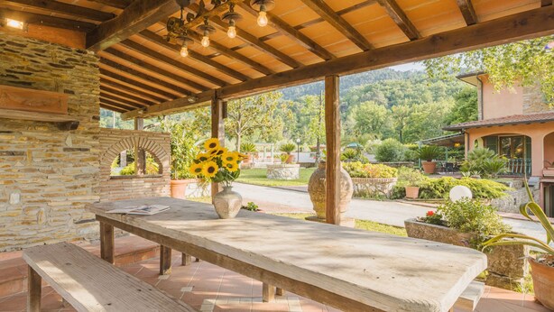 A covered outdoor seating area with stone walls, wooden beams, and a view of a lush garden and hillside. - Costacce by Interhome (Camaiore)