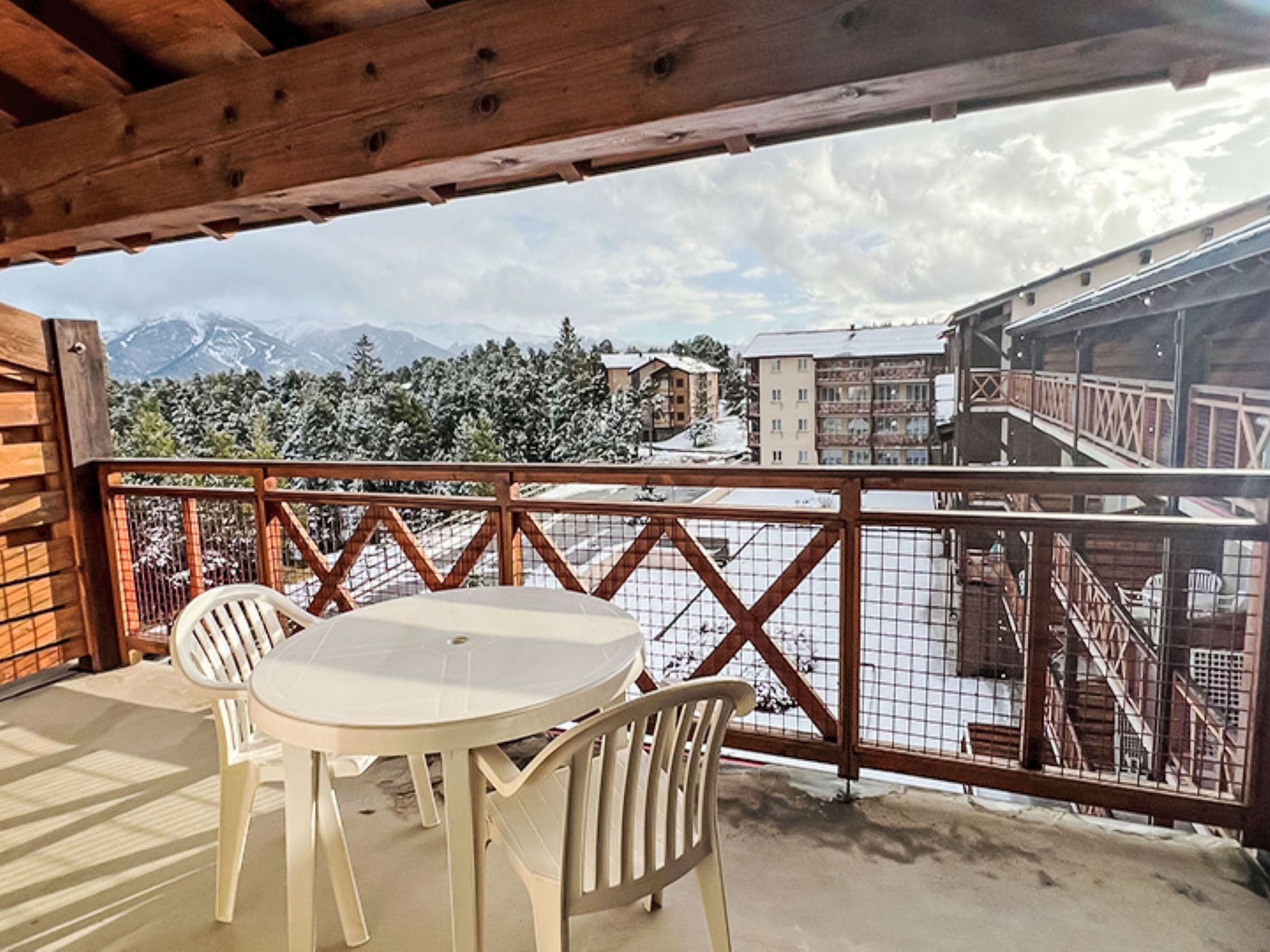 Ein überdachter Außenbereich mit Tisch und Stühlen, einem Holzgeländer und Blick auf schneebedeckte Berge und Gebäude.