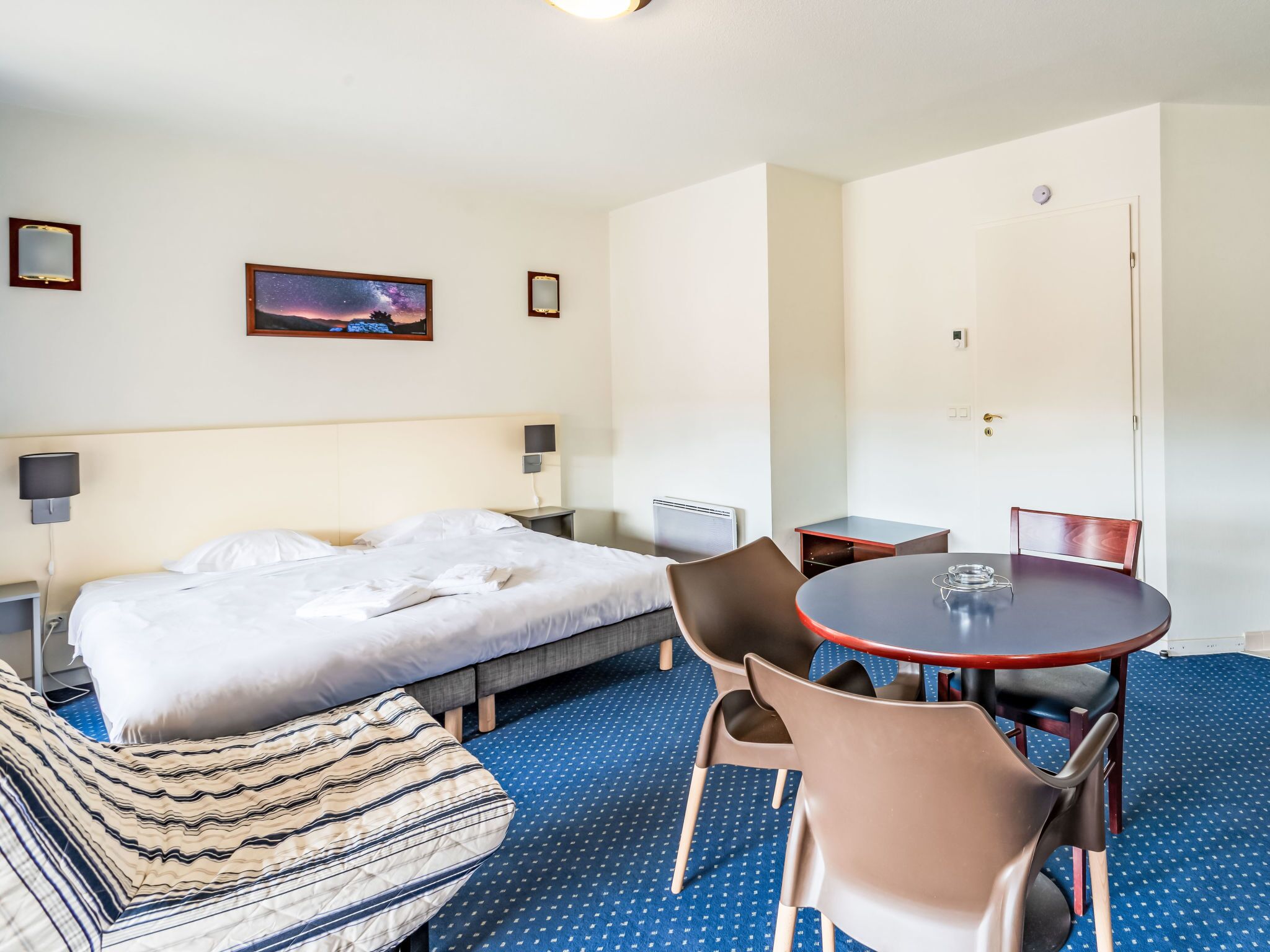 Ein Hotelzimmer mit Bett, Nachttisch, Sessel, kleinem Tisch und einem Bild an der Wand.