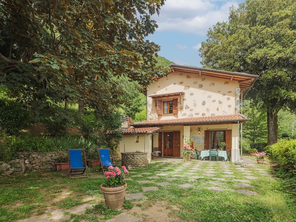 A two-story stone house with a tiled roof, a stone wall, and a paved patio with a table and chairs.