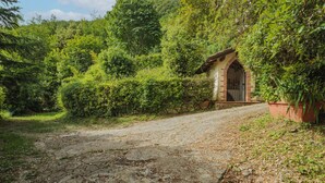 A rustic stone building with a tiled roof, surrounded by lush greenery and a gravel path leading to it.