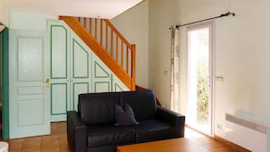 Un salón con sofá de cuero negro, una mesa de centro de madera, una escalera con barandilla de madera y una puerta que da a un balcón.