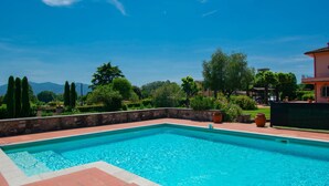 A swimming pool with a stone wall and tiled deck, surrounded by greenery and trees, with a clear sky above. - Vacation home Teresa by Interhome (Lucca)