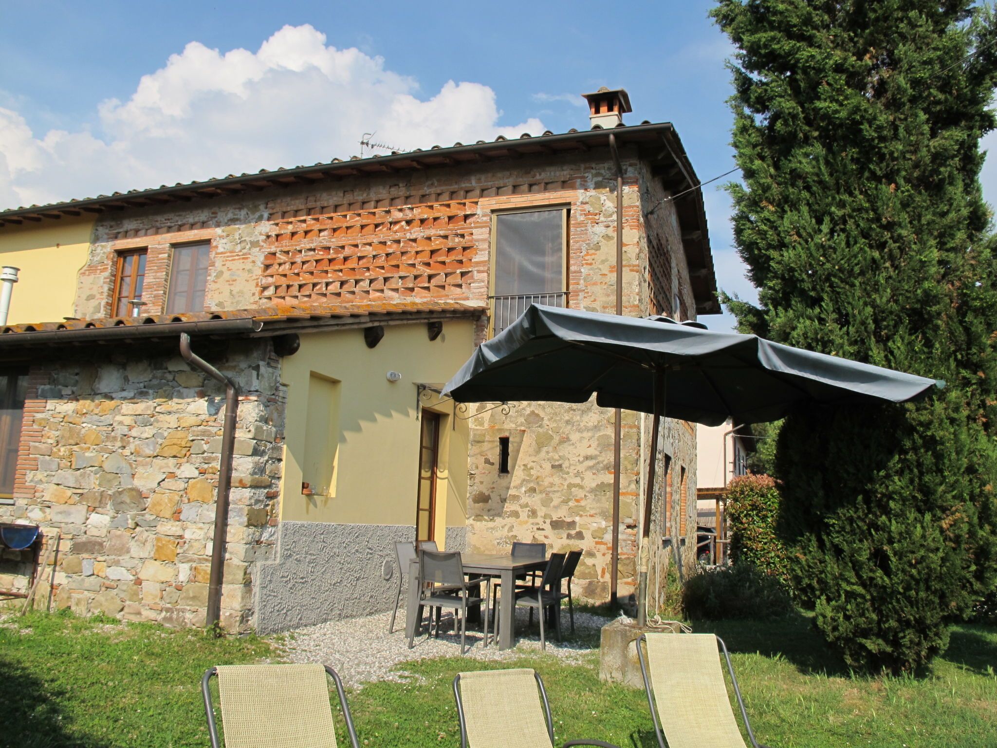 A rustic stone building with a tiled roof, outdoor seating, and a large tree nearby.