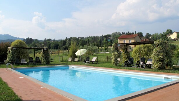 A rectangular swimming pool with a tiled deck, surrounded by lounge chairs and umbrellas, set against a backdrop of houses and greenery. - Giuseppe by Interhome (Lucca)
