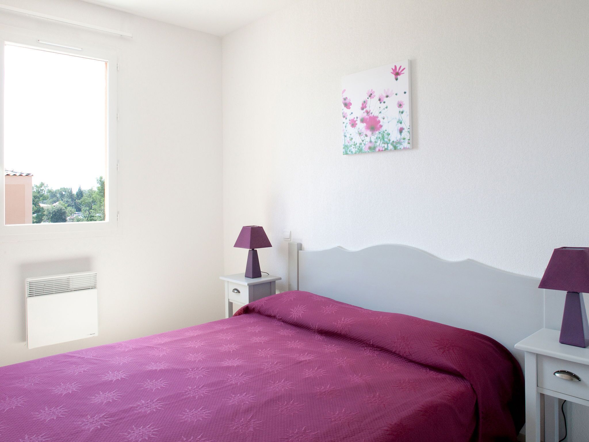 A bedroom with a bed, two bedside tables, a window, and a wall-mounted artwork.