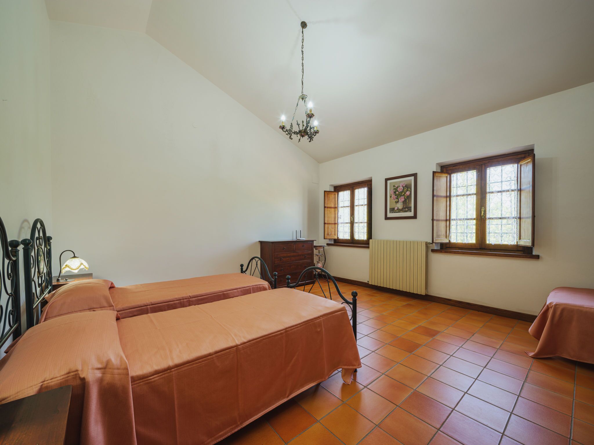 A room with two beds, a dresser, a chair, a radiator, and two windows.