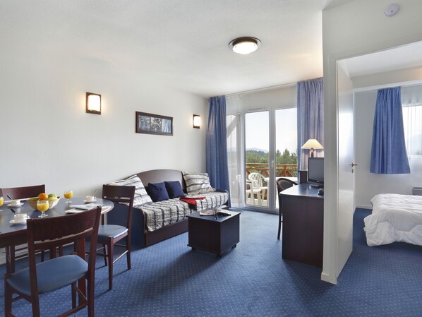 A hotel room with a bed, a sofa, a desk with a computer, a dining table with chairs, and a balcony with a view of the outdoors.