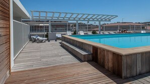 Un área de piscina en la azotea con cubierta de madera, sillones de descanso y una pérgola.