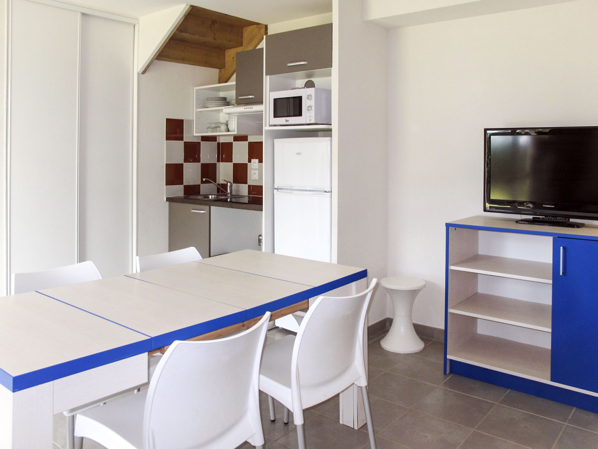 A modern kitchen with white cabinets, a microwave, and a refrigerator. There is a dining table with chairs and a TV on a blue stand.