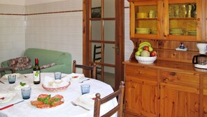 A dining area with a round table set for two, a green armchair, a wooden cabinet with glass doors, and a bowl of fruit on the cabinet.
