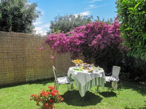 Une table ronde recouverte d’une nappe blanche, entourée de deux chaises, installée dans un jardin agrémenté d’une clôture en bambou et de plantes en fleurs.