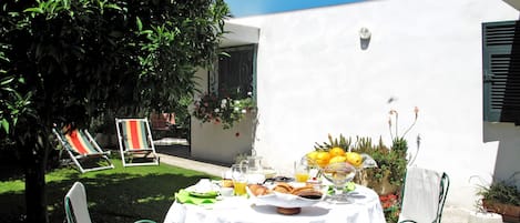 Une table dressée à l’extérieur, recouverte d’une nappe blanche, entourée de chaises vertes, et sur laquelle on trouve une variété de plats et de boissons.